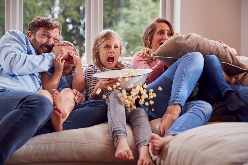 frightened family sitting on sofa at home watching horror movie on tv with popcorn